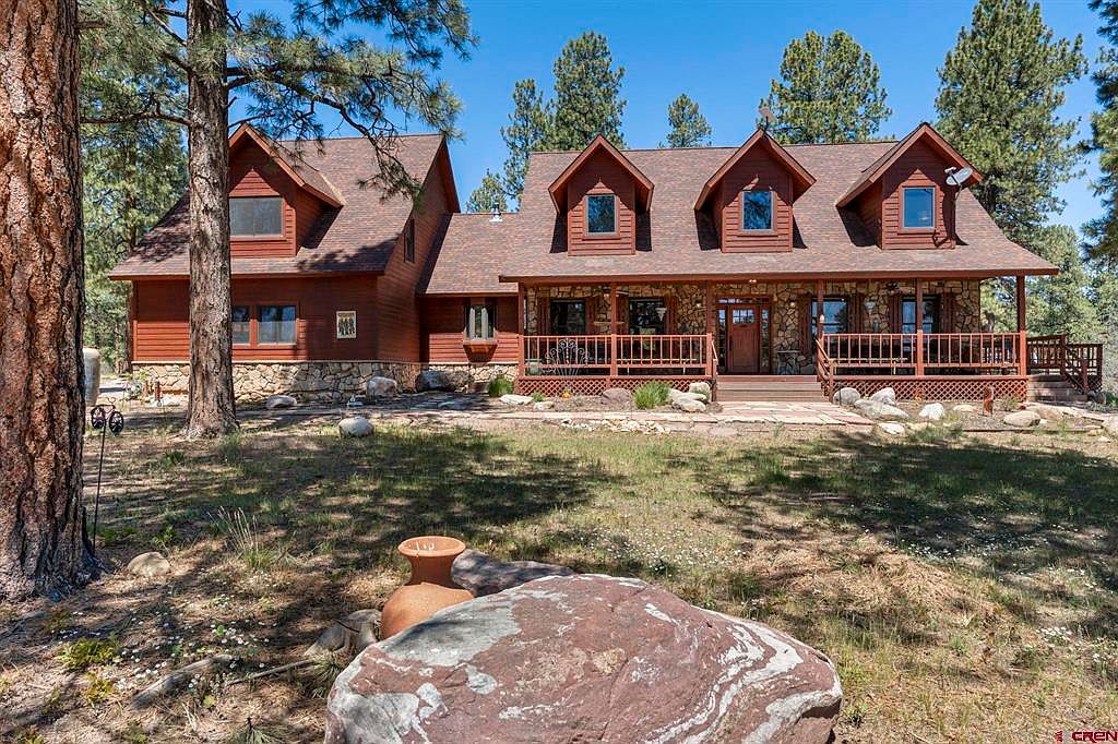 Wide front elevation of the lodge showing stone foundation, cherry door, and dormer windows under the ponderosa pines
