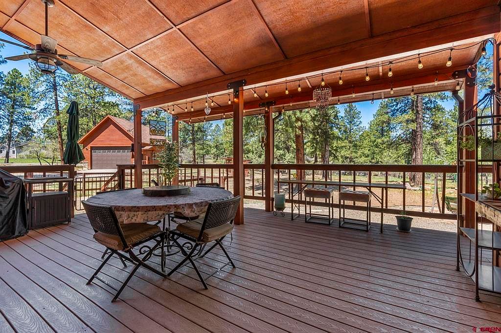 Covered back deck strung with cafe lights, a round stone-topped dining table, and the detached garage visible through the trees